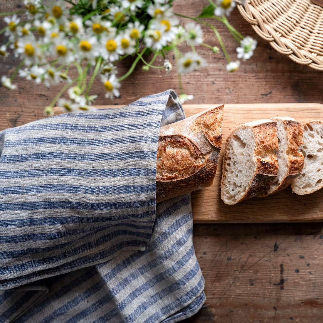 Sliced bread on a wooden cutting board with a blue and linen striped towel and flowers in the background.