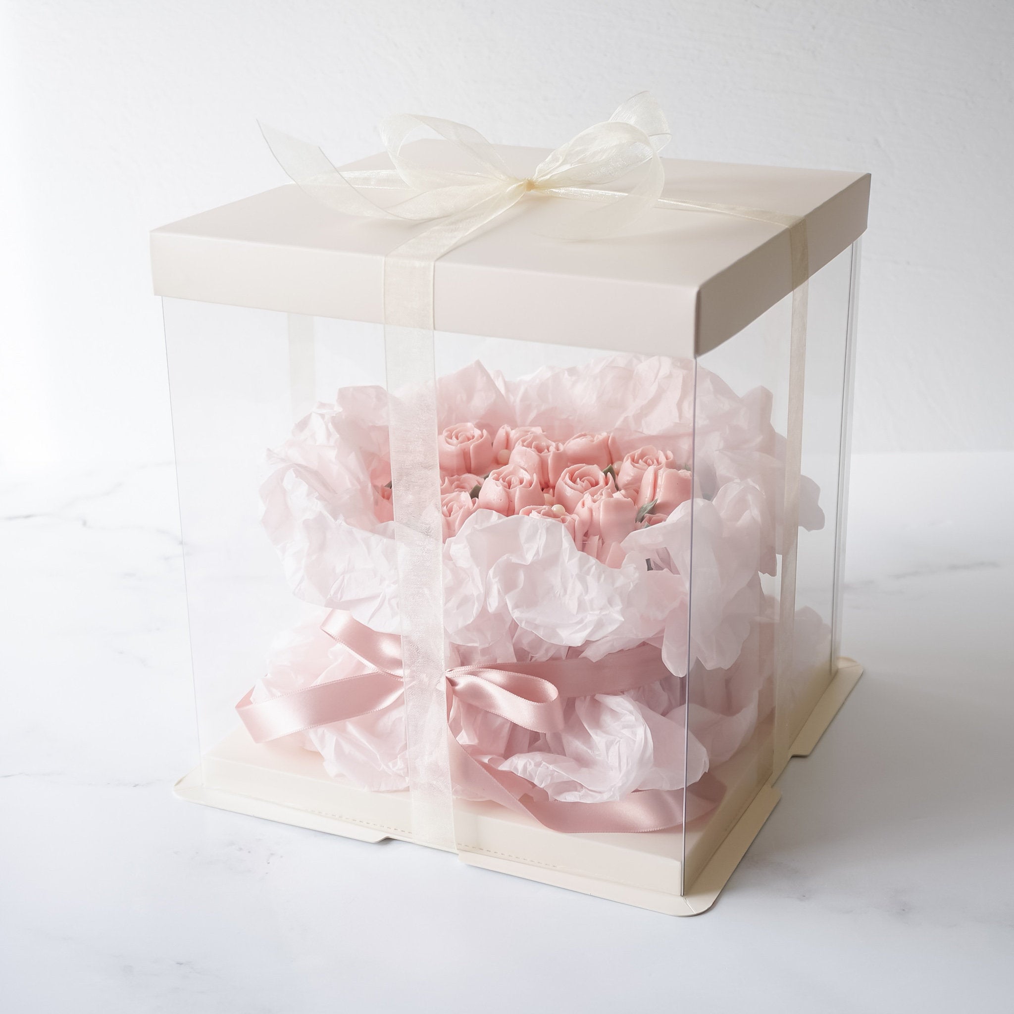 A clear plastic box with a white lid and a pink ribbon. Inside the box is a bouquet of pink roses wrapped in pink tissue paper.