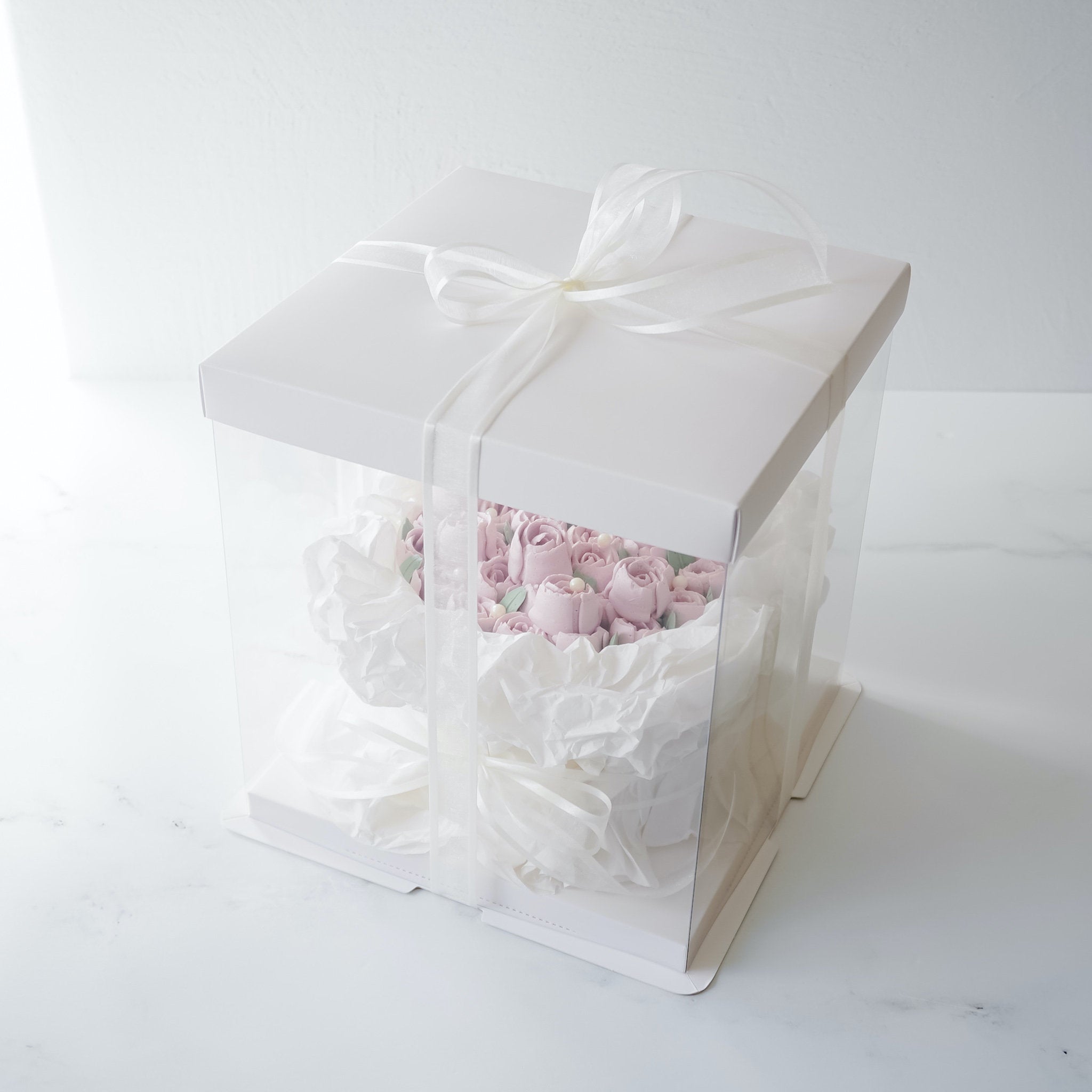 A white square box with a clear plastic window. The box is tied with a white ribbon and contains a pink flower bouquet cake made of buttercream frosting.