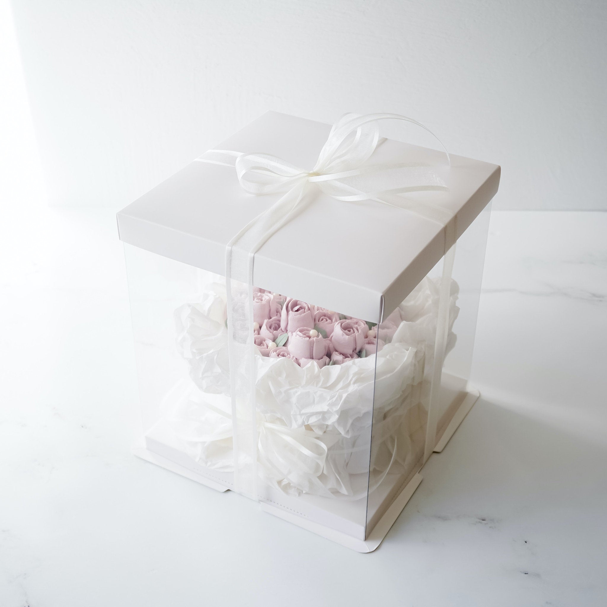 A white square box with a clear plastic window, containing a pink and white buttercream flower bouquet cake. The box is tied with a white satin ribbon.