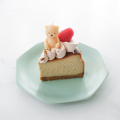 Slice of cheesecake with a teddy bear and raspberry on top, served on a green plate.