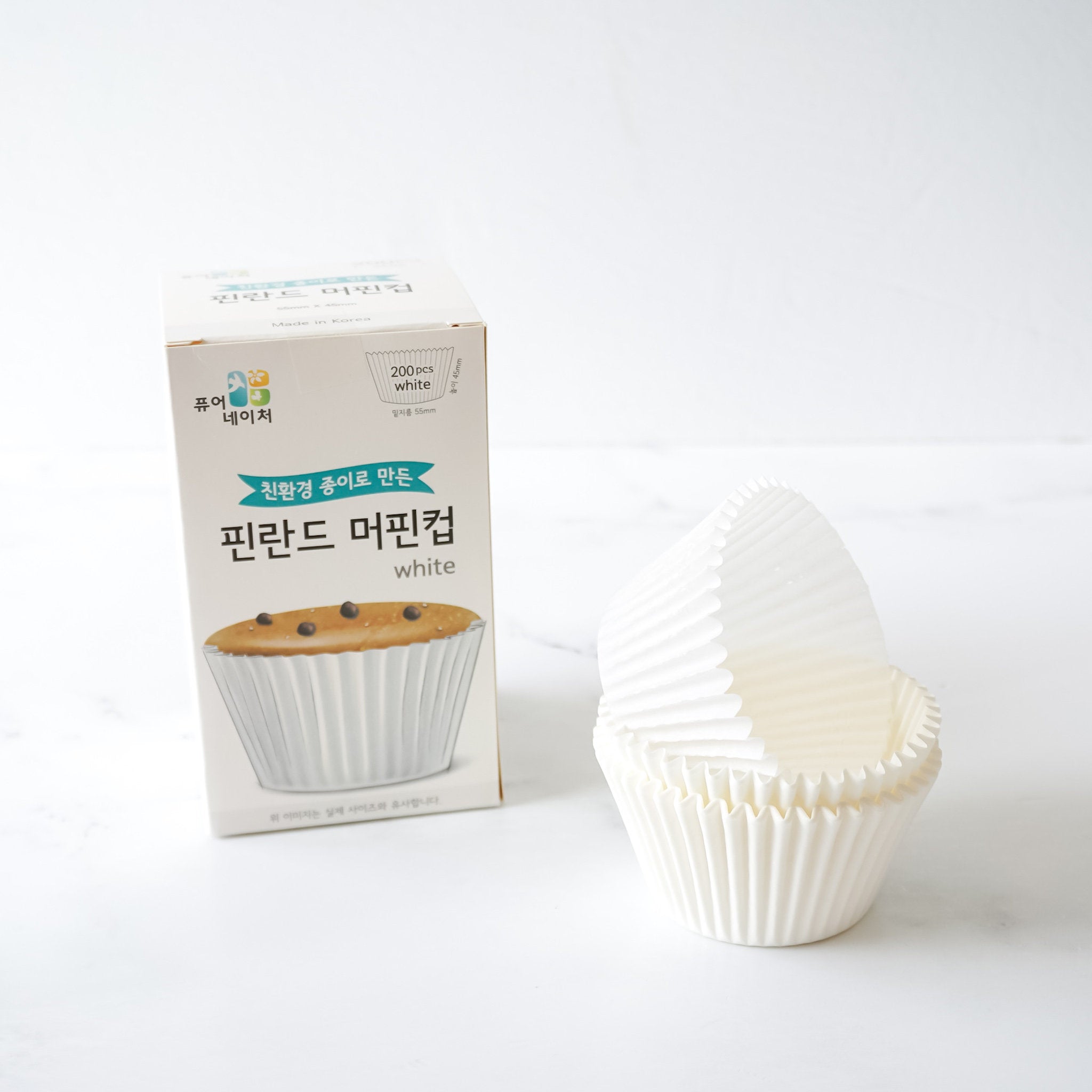A stack of white parchment paper cupcake liners. The liners are fluted and have a slight sheen. The liners are next to a box of brown parchment paper cupcake liners. 