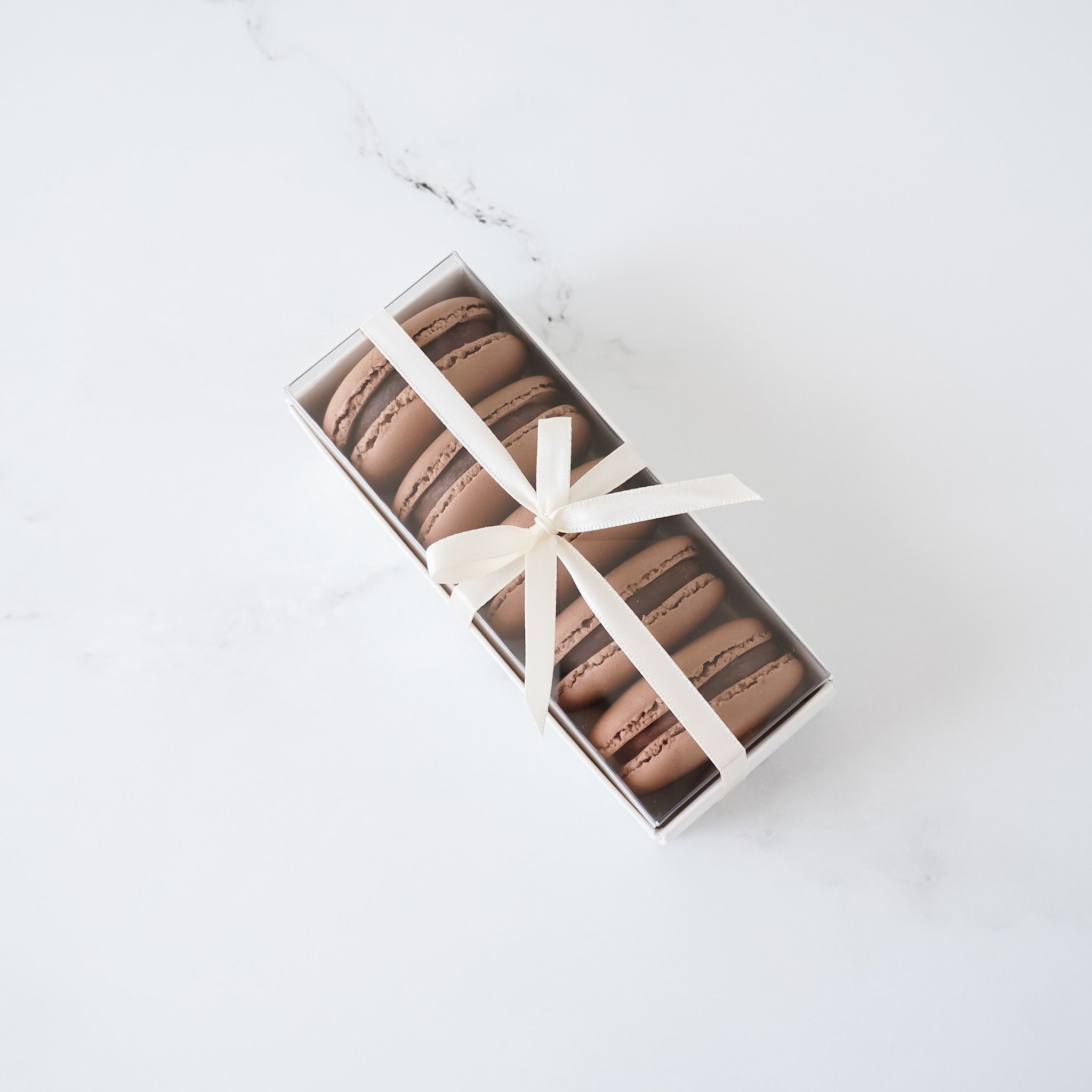 A box of chocolate macarons tied with a white ribbon. The macarons are arranged in a row and are a light brown color.