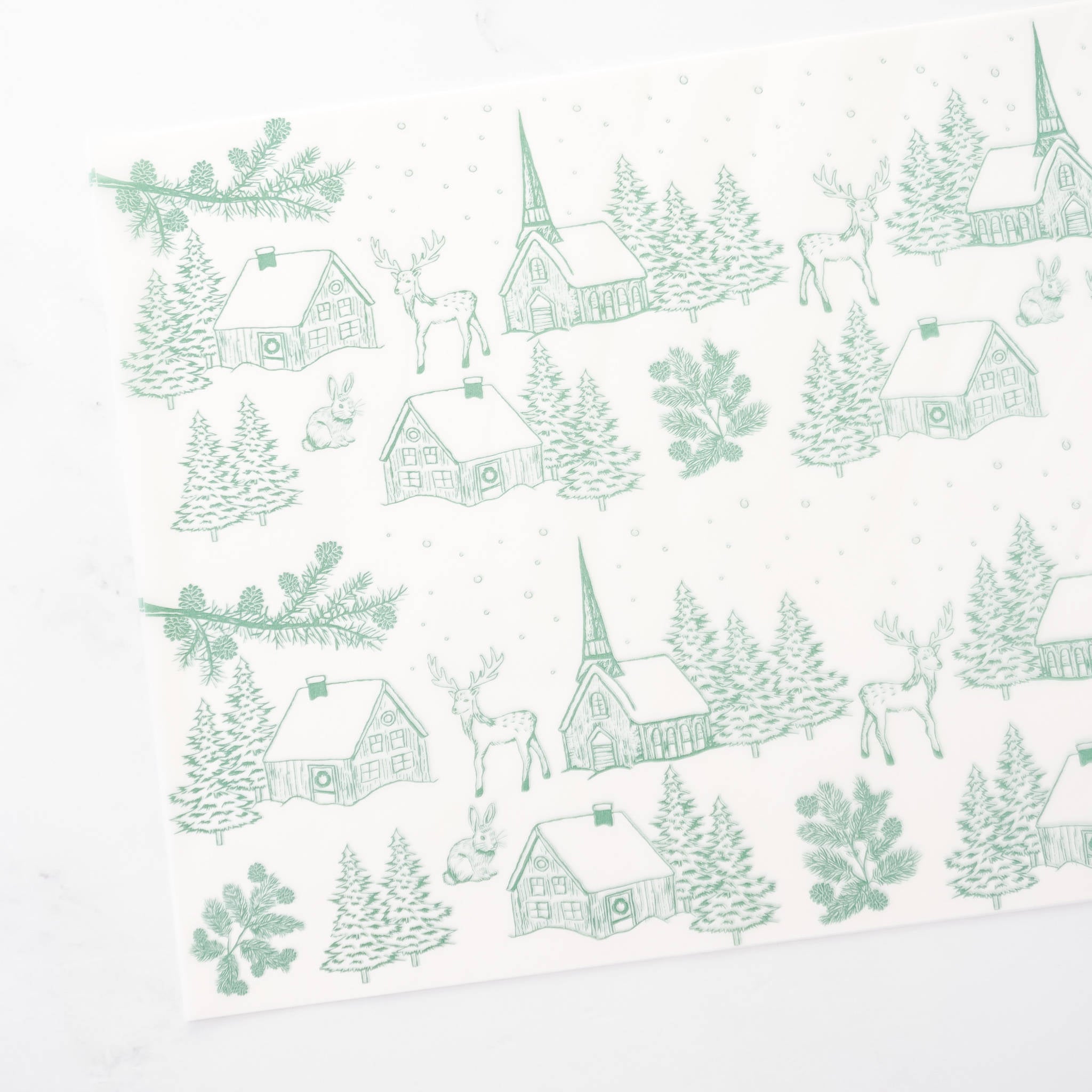 A sage green and white Christmas wrapping paper features a repeating pattern of charming illustrations. The design includes quaint houses, snow-covered trees, playful deer, and delicate pine sprigs, creating a festive winter wonderland scene.  The paper&