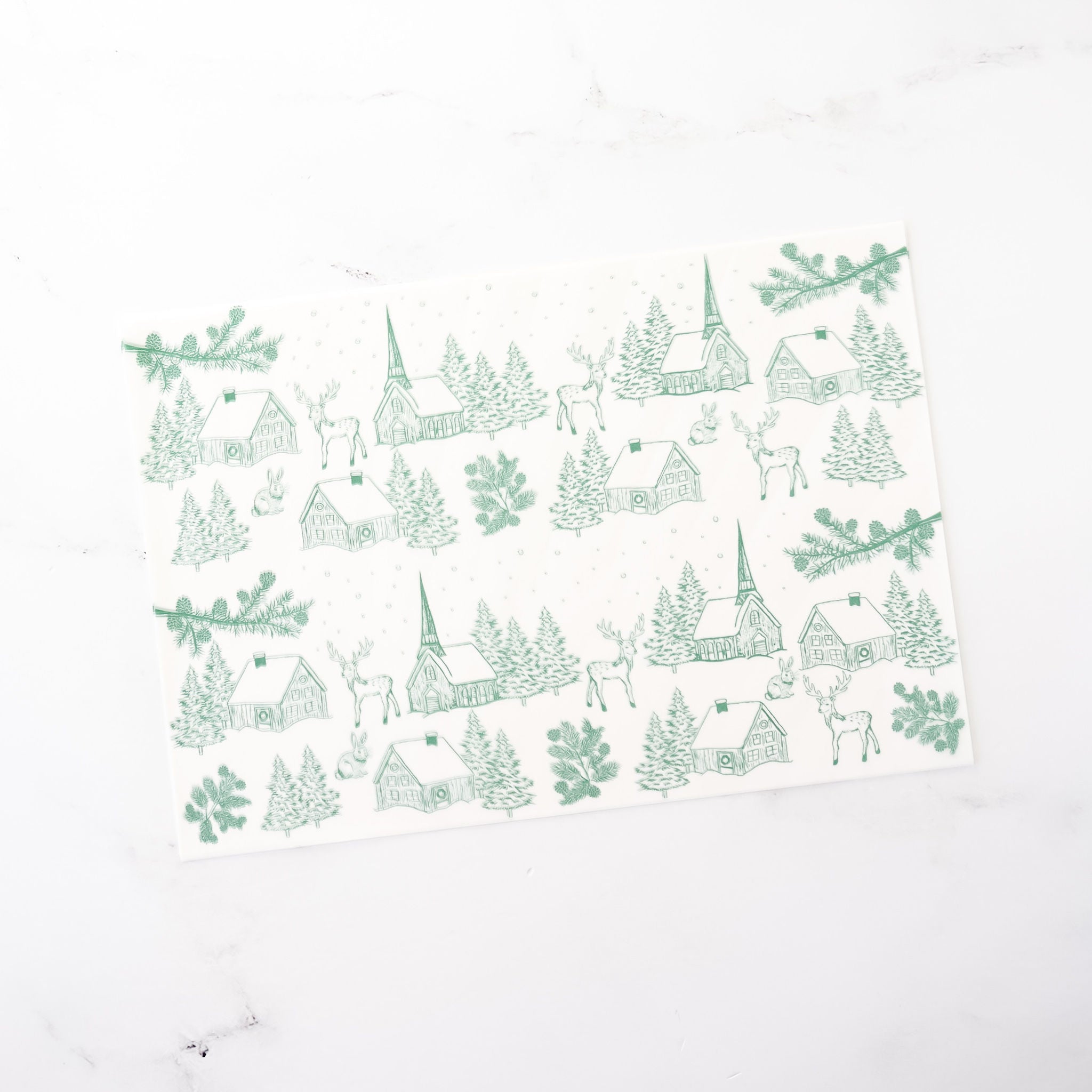 Green Christmas wrapping paper features a repeating pattern of houses, churches, deer, rabbits, and pine trees on a white background. The design is simple and elegant, perfect for holiday gift-wrapping.