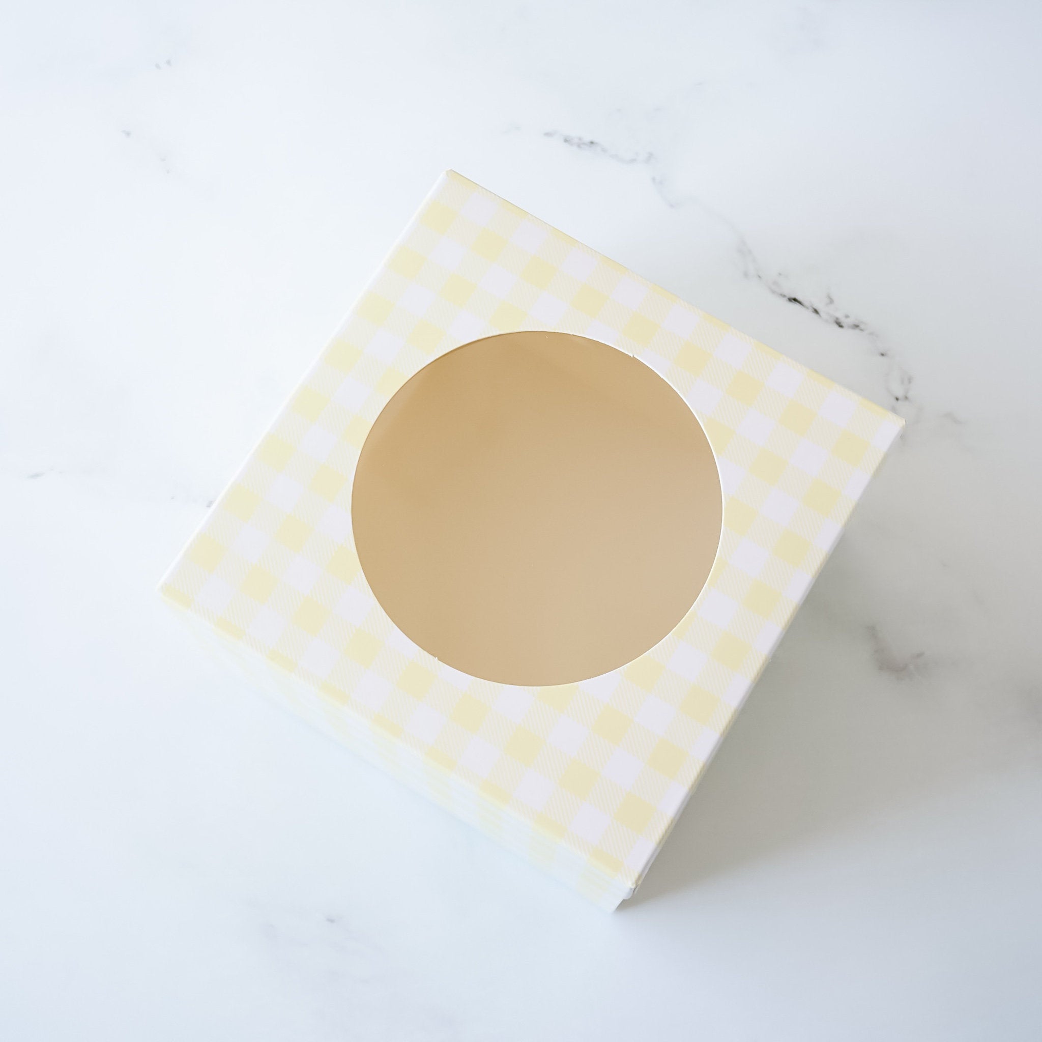 A white square box with a yellow checkered pattern and a circular cutout in the center. The box is sitting on a white marble surface.