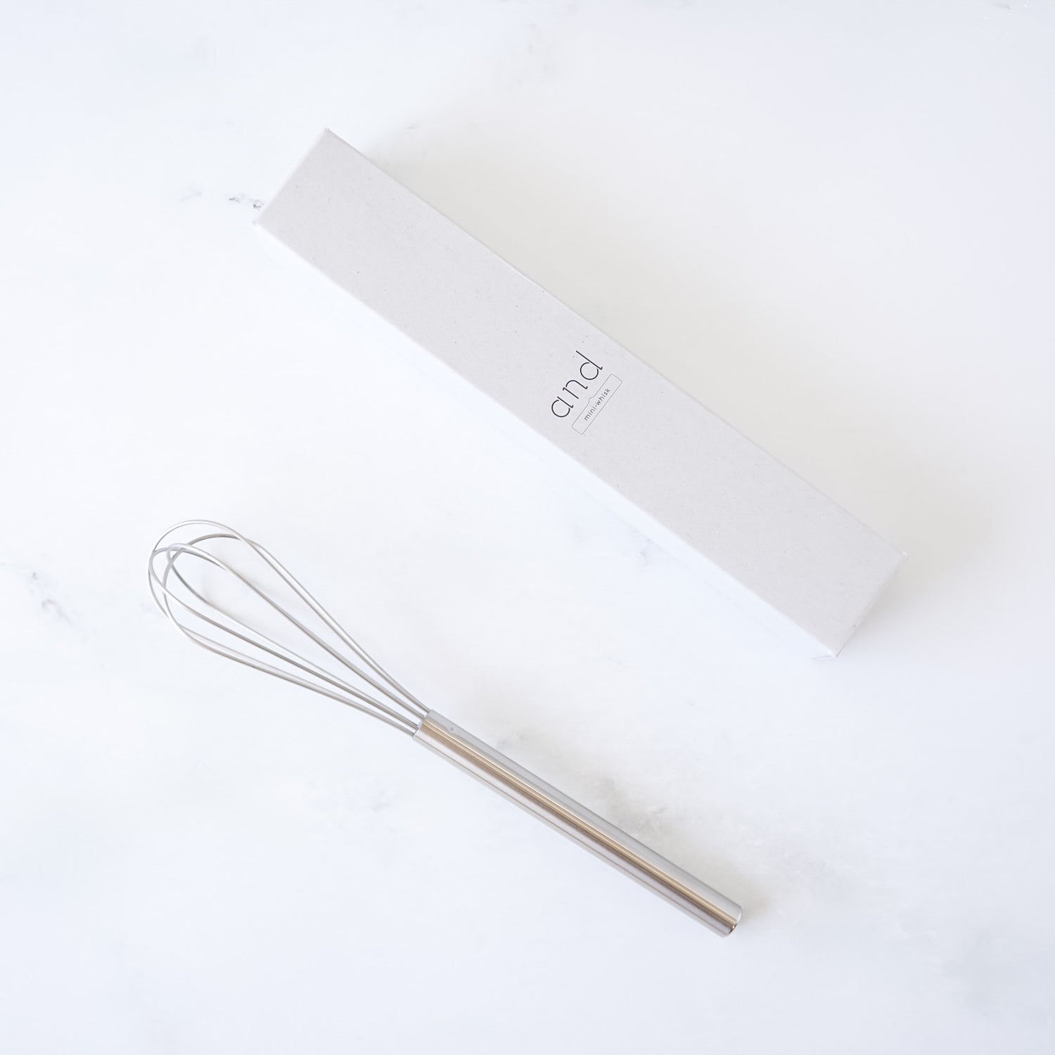 stainless steel mini whisk on a white surface with a branded box in the background