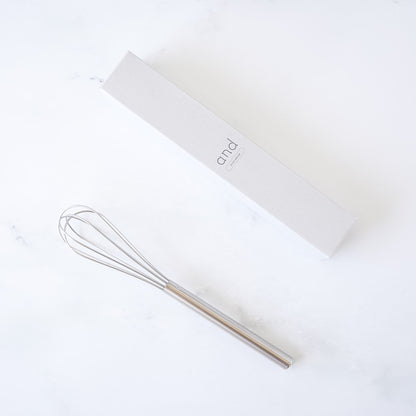 stainless steel mini whisk on a white surface with a branded box in the background