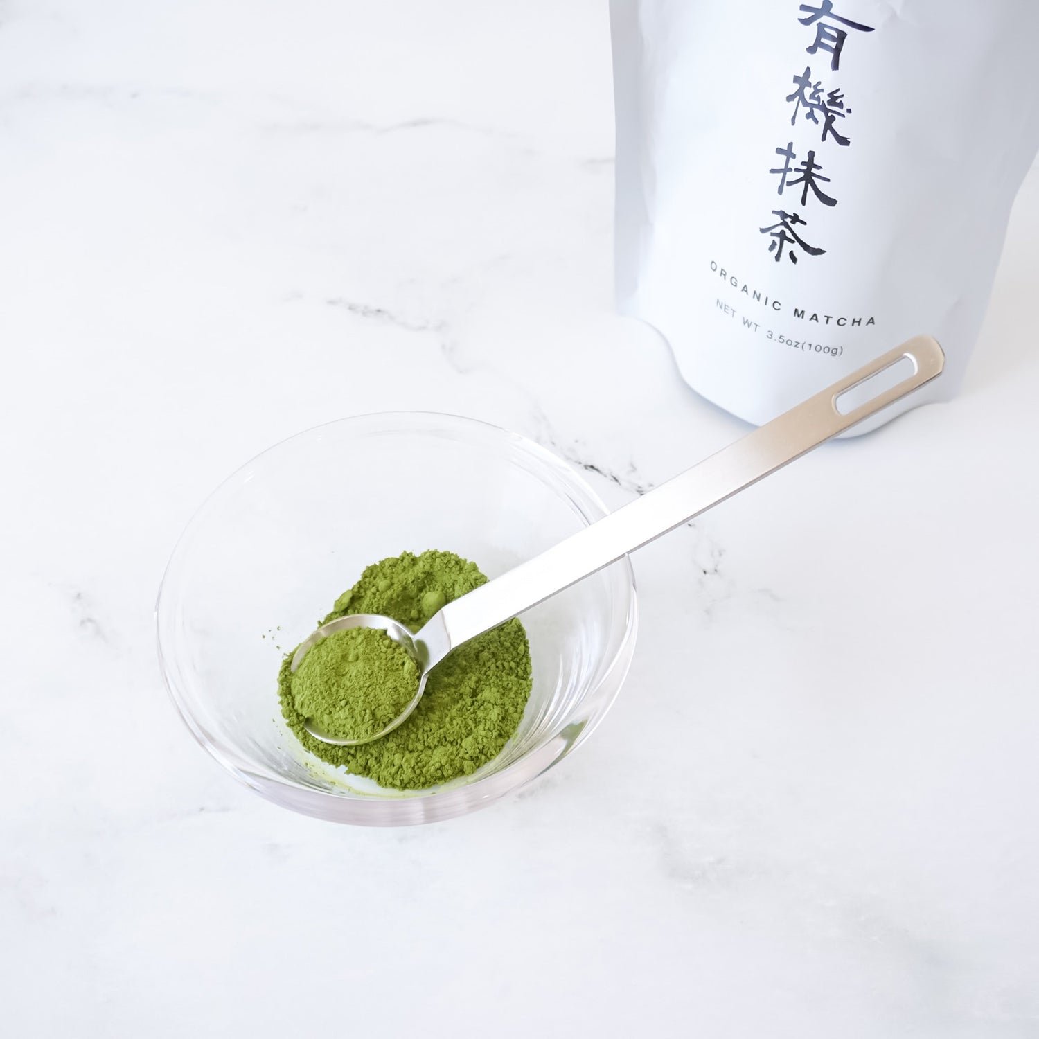 Matcha powder in a glass bowl with a teaspoon, next to a container labeled &